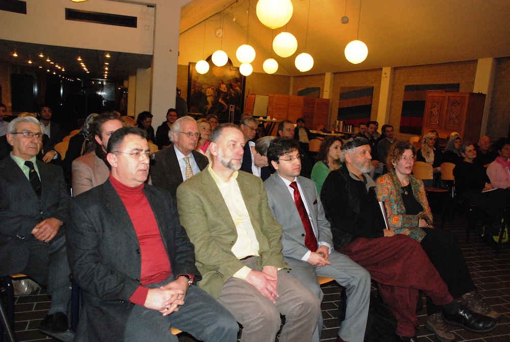 Publiek luistert in een zaal met ronde hanglampen tijdens een muziek- en poëzieavond