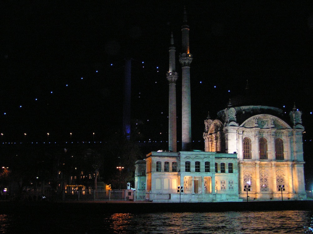 Verlichte moskee aan de Bosporus in Istanbul bij nacht, met daarachter de brug in blauw licht