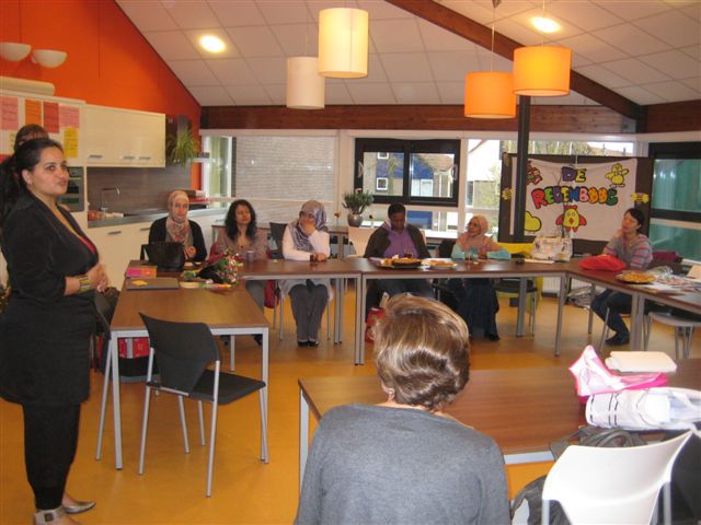 Vrouw spreekt een groep vrouwen toe aan tafels in een lokaal van een basisschool