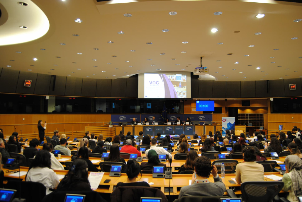 Volle vergaderzaal van het Europees Parlement met een panel op het podium en het VOC-logo op het scherm